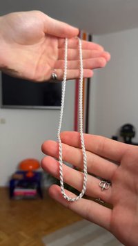 Srebrna vponka verižice, držana med dvema rokama; ima gladko teksturo in podolgovate ovalne zanke. Minimalističen dizajn brez okraskov.