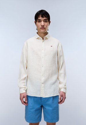 Ung mand iført en hvid langærmet skjorte og blå shorts, står foran en ensfarvet lys grå baggrund.