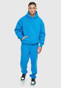 Niebieska bawełniana bluza z kapturem i kieszenią z przodu, zestawiona z dopasowanymi niebieskimi spodniami dresowymi. Czaro-białe sneakersy dopełniają outfit.