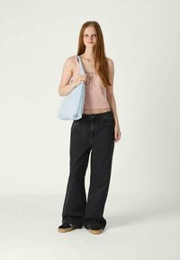 Mulher jovem com cabelo longo e ruivo, usando uma blusa rosa sem mangas, jeans largos pretos, tênis pretos, segurando uma mala de ombro azul clara.