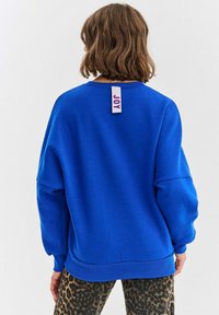 Modrá mikina s voľnými ramenami, rebrovanými manžetami a lemom. Na zadnej strane sa nachádza biely štítok s červeným a modrým textom. Kombinovaná s legínami s leopardím vzorom.