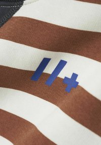 Tessuto a strisce marroni e crema con una consistenza morbida. Presenta numeri stampati in blu "11" con un accento "+". Il bordo ha una cucitura scura.