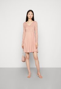 Vestido rosa com mangas longas de renda e decote em V, a alargar a partir da cintura. Usado com sandálias transparentes de salto e uma pequena bolsa rosa.