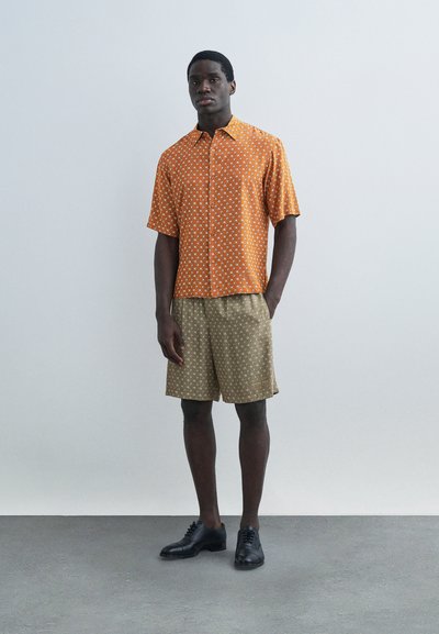 Άνδρας στέκεται φορώντας πορτοκαλί πουκάμισο με κοντά μανίκια και σχέδια, μπεζ σορτς με σχέδια και μαύρα επίσημα παπούτσια, μπροστά σε απλό γκρι φόντο.