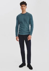 Turkusowy sweter z dzianiny o okrągłym dekolcie, długich rękawach i ściągaczach, w zestawie z ciemnymi spodniami i czarnymi butami. Prosty fason.