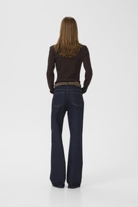 Woman with long brown hair wearing a dark long-sleeve top and dark blue wide-leg jeans, standing with hands in pockets facing away.
