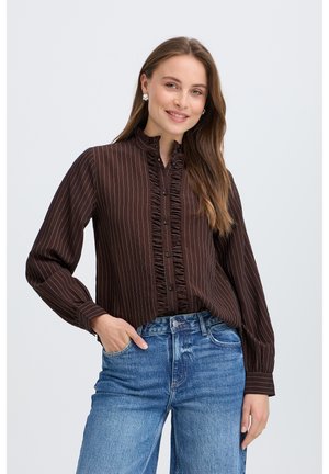 Kvinde med langt brunt hår iført en mørkebrun, stribet bluse med flæser og blå jeans, smilende med den ene hånd i lommen.