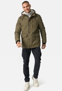 Olijfgroen jack met capuchon, knoopsluitingen en borstzakken. Gecombineerd met zwarte cargobroek en beige laarzen. Glad, duurzaam materiaal.