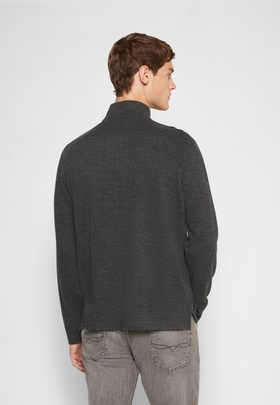 Camisa polo de manga longa cinza escuro com uma textura suave e um colarinho alto, combinada com umas calças de ganga cinza claro com bolsos traseiros distintos.