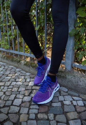 Legs in black leggings wearing purple and blue running shoes standing on cobblestone pavement next to metal fence with greenery.