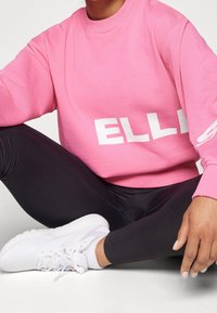 Un sweat-shirt rose avec des lettres blanches repose sur un legging noir, assorti à des chaussures de sport blanches. Le tissu semble doux avec un col rond.