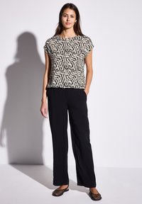 Top beige chiaro con motivo astratto nero, maniche corte; abbinato a pantaloni neri di taglio largo e scarpe nere in stile rete.