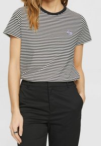 Schwarz-weiß gestreiftes T-Shirt mit kurzen Ärmeln, Rundhalsausschnitt und kleinem gestickten Logo auf der Brust. Kombiniert mit schwarzen Hosen.