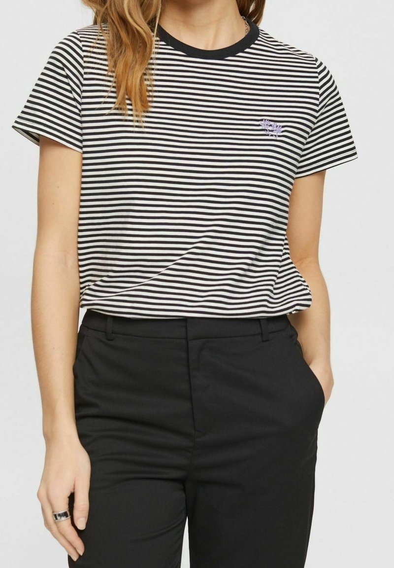 Schwarz-weiß gestreiftes T-Shirt mit kurzen Ärmeln, Rundhalsausschnitt und kleinem gestickten Logo auf der Brust. Kombiniert mit schwarzen Hosen.