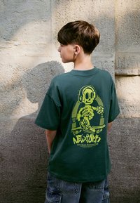 Camiseta verde de algodón con un gráfico amarillo de un esqueleto patinando con auriculares. Manga corta y corte holgado.