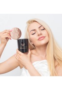 Candela nera con logo dorato e coperchio in rosa dorato, tenuta da una persona con capelli lunghi e chiari. Texture liscia e finitura lucida.