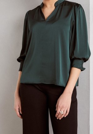 Blouse en satin vert émeraude avec un décolleté en V, des manches bouffantes et des détails aux poignets, associée à un pantalon foncé. Texture lisse, design décontracté.