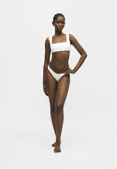 Femme grande à la peau foncée portant un maillot de bain deux pièces blanc, debout pieds nus avec une main sur la hanche, sur un fond clair uni.