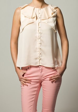 Blouse sans manches crème avec des épaules volantes et un fermoir à boutons sur le devant, associée à un pantalon ajusté rose clair. Texture de tissu lisse.
