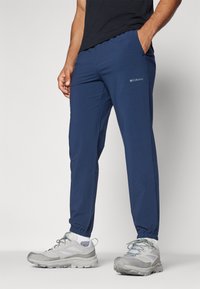 Pantalons de sport bleu marine avec ceinture élastique, poches zippées et logo Columbia. Tissu léger avec une texture lisse et coupe ajustée.