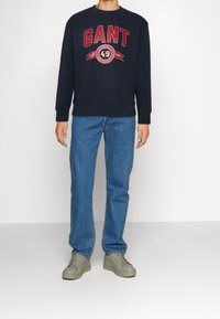 Sudadera de marina con el logo "GANT" y acentos rojos, cuello redondo, puños acanalados, combinada con jeans azul claro y zapatillas grises.