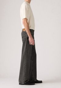 Vue latérale d’une personne portant une chemise blanche à manches courtes, un jean noir à jambes larges et des chaussures noires sur un fond uni.