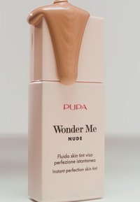 Pupa Wonder Me Nude huidtint in een rechthoekige verpakking met een lichtroze kleur, bovenop bedekt met een glanzende, bruine vloeistof die van de dop druppelt.