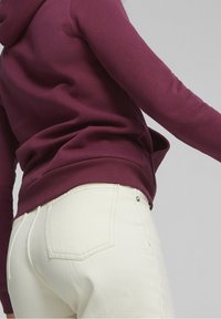 Bordeaux hætteklædning med en struktureret overflade og en kengurulomme; parret med hvide jeans med baglommer og en subtil syningsdetalje.
