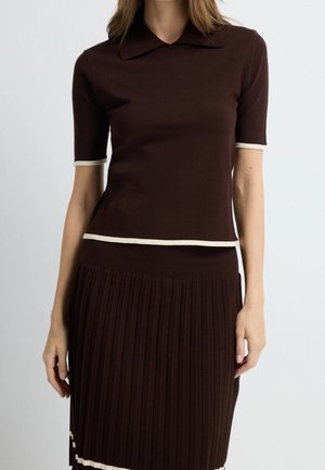 Femme portant un haut marron foncé à manches courtes avec bordure blanche et une jupe plissée assortie sur un fond clair uni.