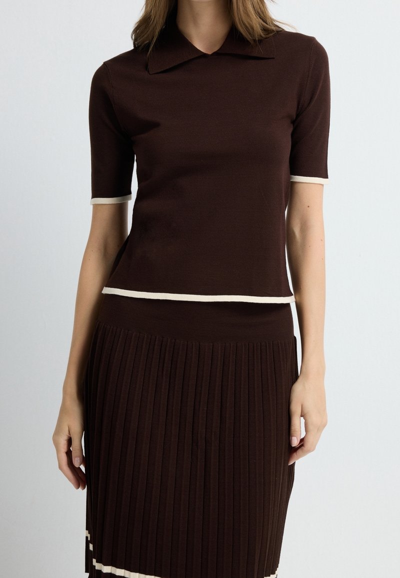 Femme portant un haut marron foncé à manches courtes avec bordure blanche et une jupe plissée assortie sur un fond clair uni.
