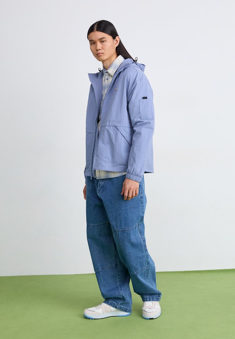 Cazadora ligera de color azul claro con capucha, que cuenta con bolsillos frontales y puños elásticos, combinada con unos vaqueros azules de corte holgado y zapatillas blancas.