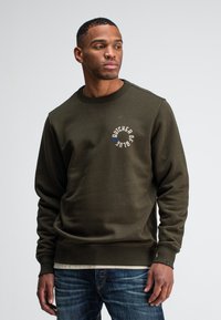 Olijfgroen crewneck sweatshirt van zachte stof, met een cirkelvormig logo waarop "BUTCHER OF BLEU" staat in beige en blauwe accenten.