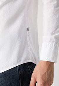 Camisa blanca de manga larga con una textura suave, que presenta un dobladillo cosido, puños abotonados y una pequeña etiqueta con el logo en la costura lateral.