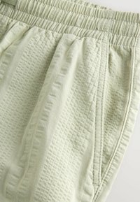 Gros plan sur un pantalon en tissu vert clair texturé avec une ceinture élastique et des détails de couture sur la couture latérale.