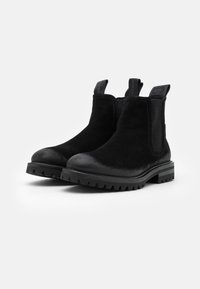 Paire de bottines Chelsea en daim noir avec languettes de traction, panneaux latéraux élastiques et semelles crantées sur fond blanc.