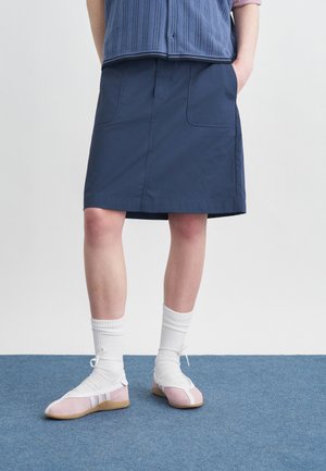 Person iført navyblåt skørt, hvide sokker og lyserøde snøresko, der står på blåt tæppe med hænderne i lommerne.