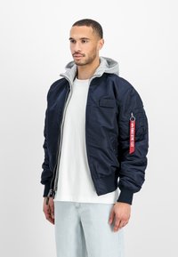 Hombre con chaqueta bomber azul marino y capa interior con capucha gris, camiseta blanca y jeans azul claro, de pie frente a un fondo blanco.