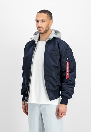 Man som bär en marinblå bomberjacka med grå huva inuti, vit t-shirt och ljusblå jeans, stående mot en vit bakgrund.