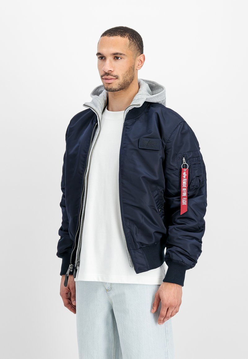 Hombre con chaqueta bomber azul marino y capa interior con capucha gris, camiseta blanca y jeans azul claro, de pie frente a un fondo blanco.