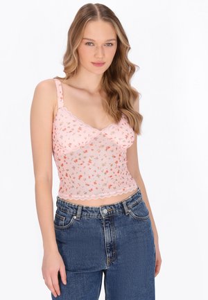 Tânără cu păr lung și ondulat, purtând un maiou roz cu motiv floral și blugi albastri cu talie înaltă, stând pe un fundal alb.