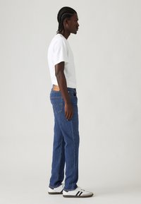 Hombre con el cabello trenzado de pie de lado, vistiendo una camiseta blanca, jeans azules y zapatillas blancas con rayas negras sobre un fondo liso.