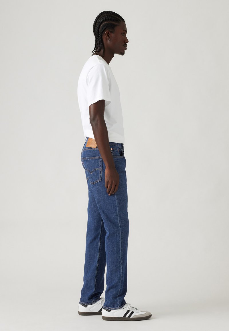 Mann mit geflochtenem Haar steht seitlich und trägt ein weißes T-Shirt, eine blaue Jeans und weiße Sneakers mit schwarzen Streifen vor einem einfarbigen Hintergrund.