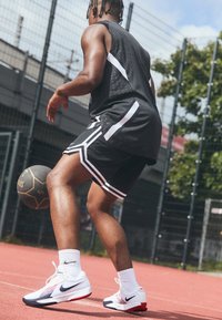 Sort atletisk tanktop med hvide accenter, sorte shorts med hvide striber og hvide atletiske sko med røde og marineblå detaljer, på en basketballbane.