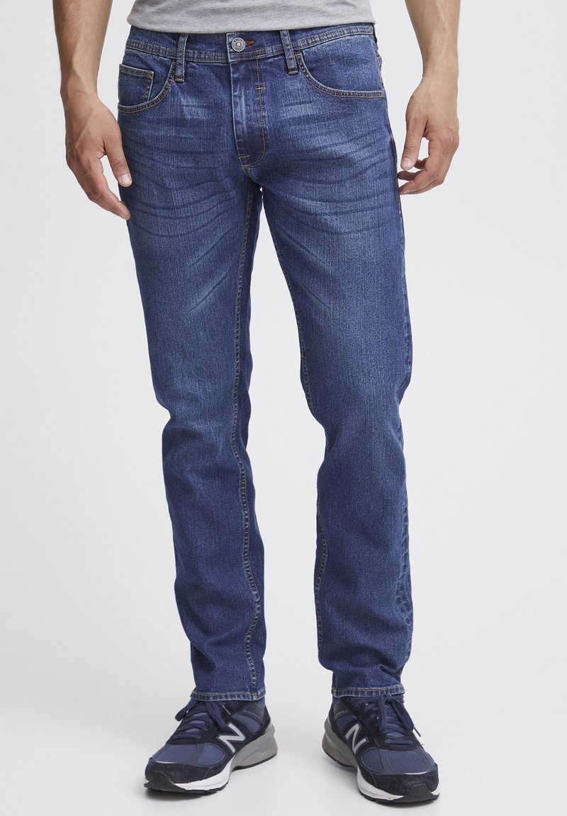 Jeans de mezclilla azul oscuro con corte recto, cinco bolsillos y un ligero desgaste. Combinados con zapatillas negras.