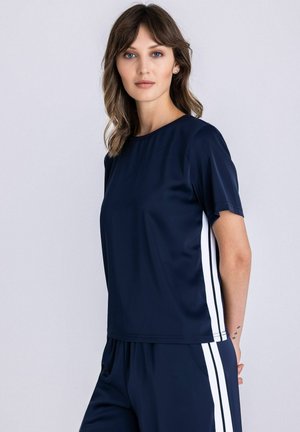 Vrouw met golvend bruin haar, gekleed in een marineblauw shirt met korte mouwen en een broek met witte verticale strepen aan de zijkanten, staande in zijaanzicht.