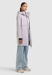 Kobieta ubrana w jasnofioletową pelerynę przeciwdeszczową z kapturem, białą bluzkę, jasnoniebieskie szerokie dżinsy i beżowe trampki, stojąca bokiem.