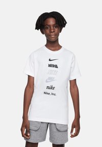 Camiseta de algodón blanca con múltiples logotipos de Nike en impresión negra y gris. Cuello redondo, mangas cortas, corte relajado. Combinada con shorts grises.
