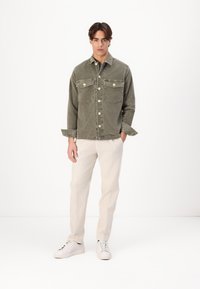 Olijf groene corduroy jas met twee borstzakken, knopenluiting en opgerolde mouwen, gecombineerd met licht beige broeken en witte sneakers.