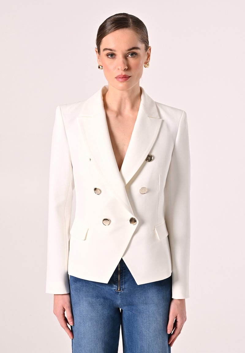 Blazer blanc à double boutonnage avec revers, boutons argentés et poches latérales, associé à un jean en denim bleu. Texture lisse en tout lieu.