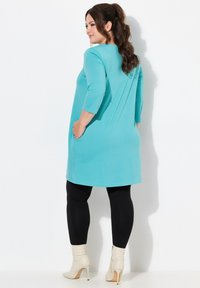 Turquoise tuniek met ¾ mouwen, voorzien van zijzakken; gecombineerd met zwarte leggings en witte enkelboots. Zachte, rekbare stof.